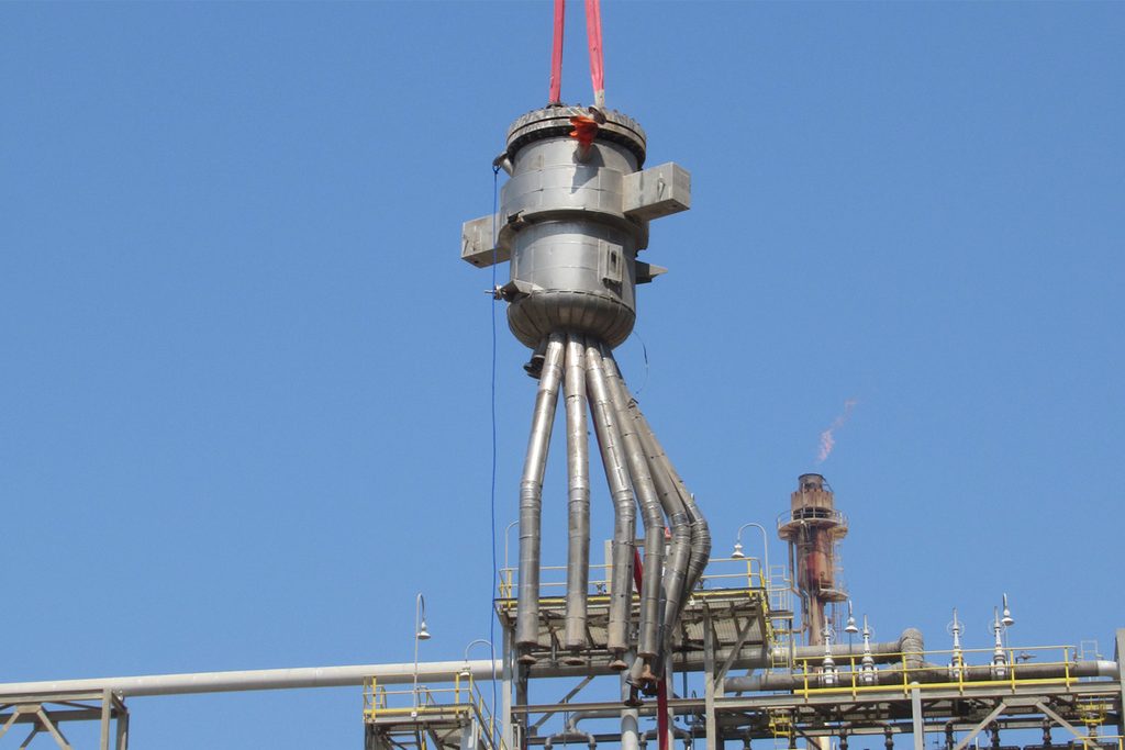 A CCR reactor component with catalyst transfer pipes being lifted by crane above the refinery structure.